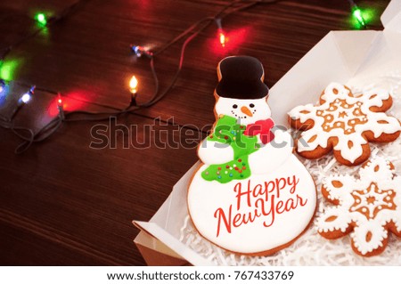 Christmas or new year gingerbread snowman and snowflakes in box on a dark table with light garlands. Concept of gift. Picture for a confectionery catalog. Invitation or greeting card with copy space.