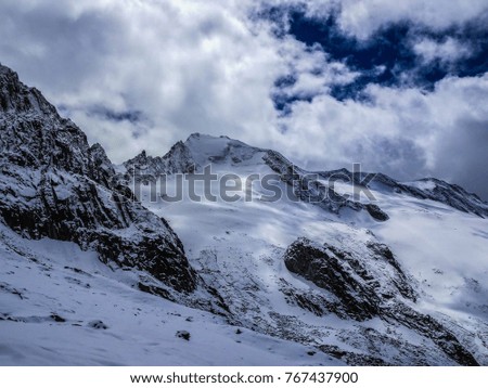 Ice covered mountain peak surrounded with ice, snow and rock and steep walls and ridges falling down from the top below a cloudy sky with a "blue window"