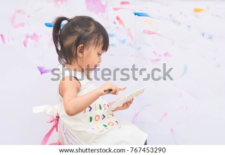 Cute asian little girl is sitting and talking something, she is holding the brush and color palette for painting the color to the white paper, concept of art and education for kid.