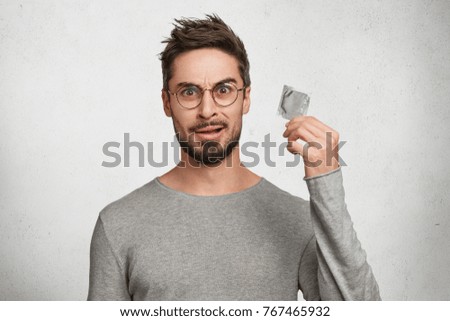 Contraception and healthy lifestyle concept. Male youngster with appealing look, holds condom, has puzzled expression as going to use it firstly. Bearded guy keeps contraceptive, advertises.
