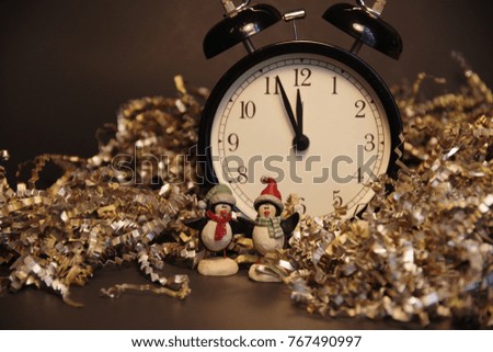 Penguins figurines with hats and scarfs in front of black alarm clock showing minutes to midnight surrounded with wrinkled gold and silver decoration paper