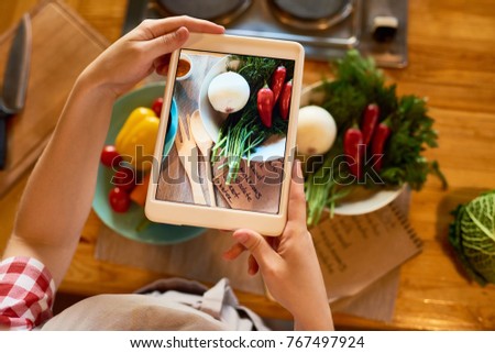 Close-up shot of female food blogger taking picture of ingredients necessary for preparing healthy dish on digital tablet while standing at kitchen table