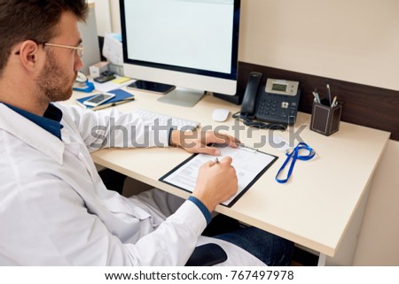 Portrait of professional physicist  sitting at desk in office and writing work notes in patients form, copy space Royalty-Free Stock Photo #767497978