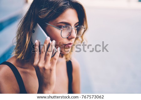 Serious young woman in trendy eyewear concentrated on information getting during phone talk with customer support operator, thoughtful hipster girl calling to friend standing near publicity area Royalty-Free Stock Photo #767573062