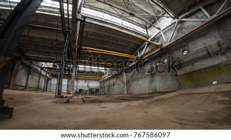Empty abandoned plant. Floor is wet. Main constructions are made of concrete. It has yellow bridge crane in a middle. Some metal blue columns stay in left side. Fish eye picture.