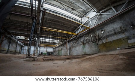 Empty abandoned plant. Floor is wet. Main constructions are made of concrete. It has yellow bridge crane in a middle. Some metal blue columns stay in left side. Fish eye picture.