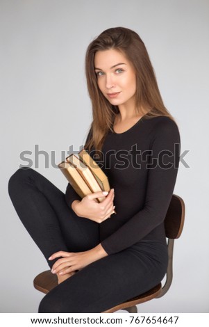 Young beautiful brunette woman sitting on a chair in neutral casual black clothes on a grey background, holding books in her hands. She is a writer