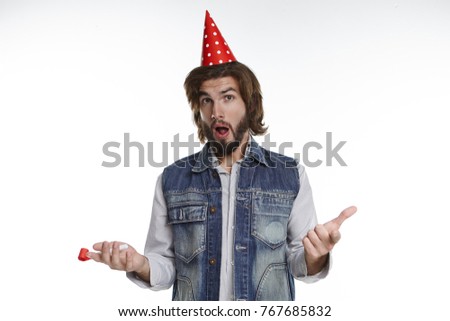 Horizontal shot of emotional young bearded man wearing stylish clothes and red holiday cap opening mouth in astonishment, shocked with surprise party thrown by colleagues to celebrate his promotion