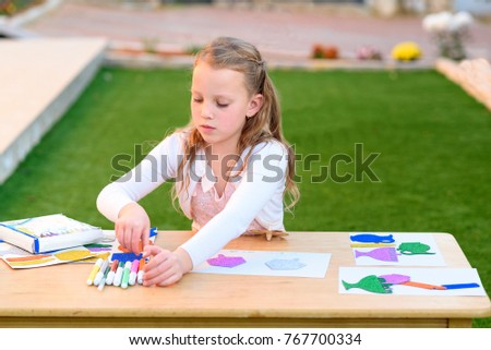 Llittle girl is cutting color paper, draw and  making handmade Hanukkah decorations: sticking menorah, candles, spinning top. Young  child doing handicrafts outdoor.
