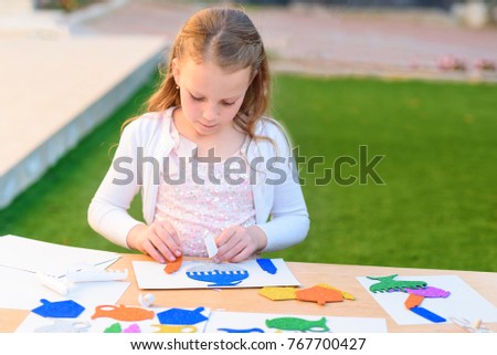 Llittle girl is cutting color paper, and  making handmade Hanukkah decorations: sticking menorah, candles, spinning top. Young  child doing handicrafts outdoor.
