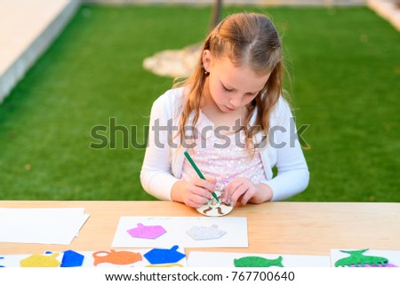 Little girl is cutting color paper, and  making handmade Hanukkah decorations: sticking menorah, candles, spinning top. Young  child doing handicrafts outdoor.
