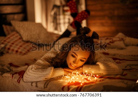 Beautiful young girl in a knitted vintage sweater with festive lights on a bed on Christmas Eve