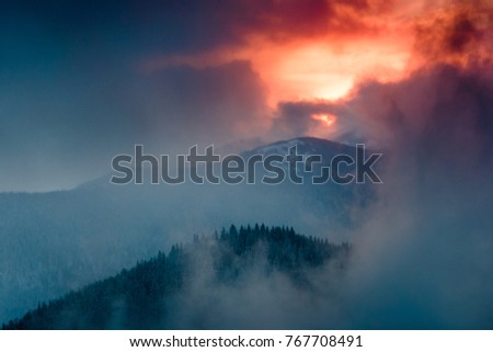 Landscape of dramatic sunset in the winter mountain. Wooded hills covered with snow, fog rising from valleys, colorful cloudy sky - this is impressive picture.