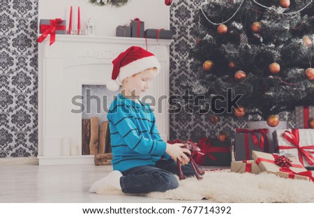Cute happy boy in santa hat holding toy. Christmas present on holiday morning in beautiful room. Male child got Xmas gift near decorated fir tree and fireplace. Winter holidays concept