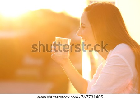Side view portrait of a happy lady holding a glass of water outside in a balcony at sunset