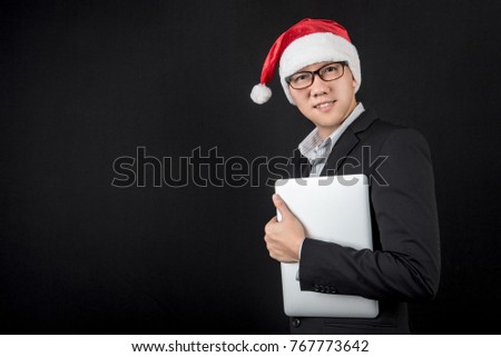 Young Asian businessman wearing suit and santa hat holding laptop computer on black background, male entrepreneur in christmas holiday and new year party concepts