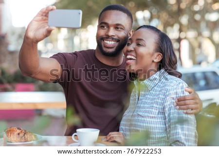 Laughing young African couple taking a selfie together at a sidewalk cafe table while out on a date 