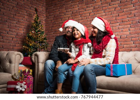 Cute little Indian/Asian Girl celebrating Christmas with Grandparents while sitting over sofa and wearing Santa Hat and with lots of gifts 