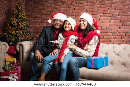 Cute little Indian/Asian Girl celebrating Christmas with Grandparents while sitting over sofa and wearing Santa Hat and with lots of gifts 