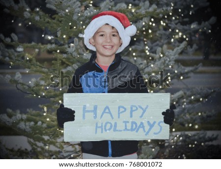 Young boy wishing you a Happy Holidays holding sign and wearing Santa clause hat with Christmas tree background lit up