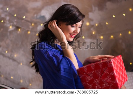 A young woman holds a gift near the Christmas decoration. The girl smiles sitting on a bed with a gift for Christmas