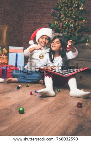 Cute little Indian/asian kids celebrating christmas while reading story book, wearing Santa Hat with lots of gifts in the background at home