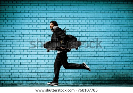 Smiling hipster dreadlock skater with headphones running in a suit near the blue wall on the street.