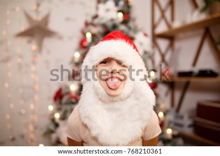 Photo of girl in Santa hat with beard showing tongue against background of New Year's decorations