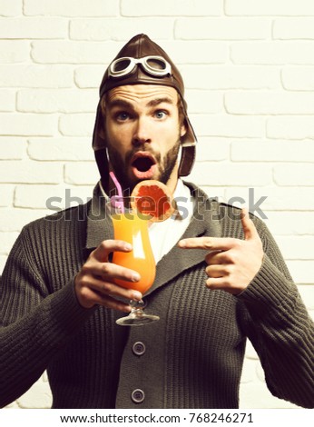handsome bearded aviator man with long beard on surprised face holding glass of alcoholic cocktail in gray knitted sweater with hat and glasses on white brick wall background