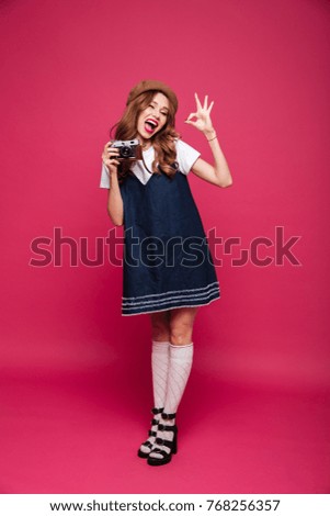 Cheerful carefree lady in blue dress showing ok gesture while holding retro camera isolated over pink