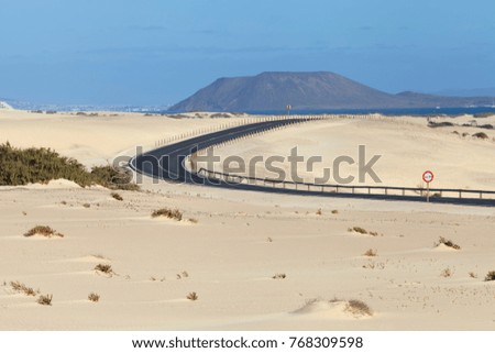 Coastline curved road through sandy dunes towards the sea, with volcanic mountains on the horizon .