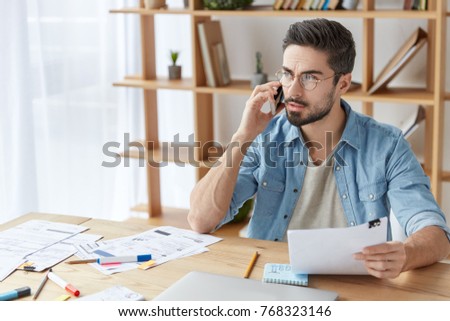 Portrait of handsome bearded young fashionable male has phone conversation, holds documents, sits at wooden table. Stylish hipster male tries to solve financial problems, sits over office interior
