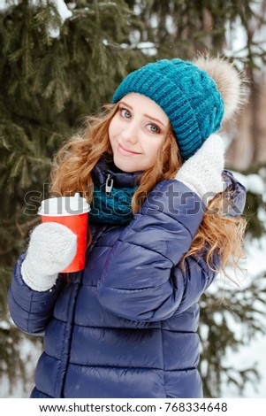 winter portrait of the lovely blue-eyed girl of the blonde with a voluminous curly hair in a blue down-padded coat against the background of snow-covered fir-trees.The girl holds in hand a red glass w