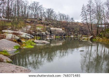 Small river floating on stone canyon among the granite rocks and the park on the background of the autumn landscape with trees without leaves and gray rainy sky. Granite Canyon of river Uzh
