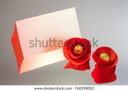 Empty blank of greeting card with red artificial flowers made of cutting paper and chocolate candy on metallic paper background with reflection and colored lighting usage.