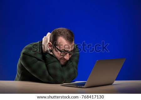 Emotional man in glasses sits at a table with a laptop and posing on a blue background