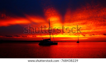 A striking inspirational Crimson altostratus cloud sunrise sky seascape with colorful bold sunbeams yacht silhouettes and glowing ocean water reflections. Australia.