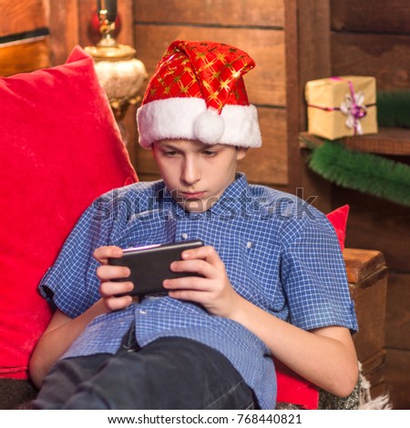 A teenager in a blue checkered shirt, in a Santa Claus hat, sits in the New Year's corner, holds his phone in his hands, and reads or plays carefully. Square picture.