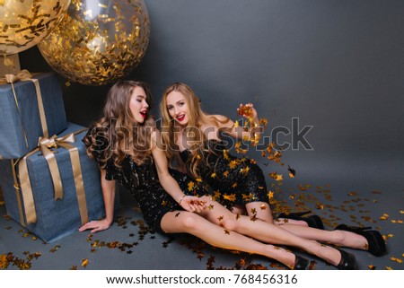 Blonde girl sitting on the floor with friend and throwing out golden confetti on dark background. Stylish ladies in black dresses lying beside presents and balloons and joking.