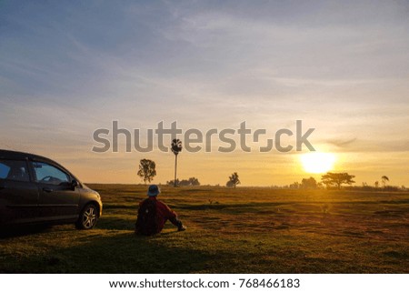 A traveling man is relaxing with beautiful sunset over field in front of him.