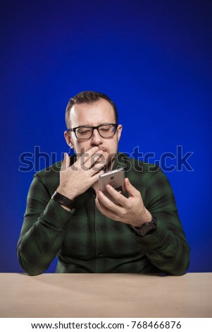 Emotional man in glasses sits at a table with a laptop, holds a phone in hands, posing on a blue background