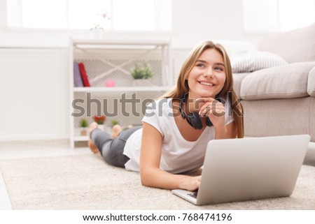 Cute smiling blonde freelancer girl working on laptop. Young woman in casual lying on the floor in light appartment.