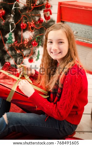 Beautiful teenage girl opening her christmas present in red clothes near the christmas tree with red decorations.