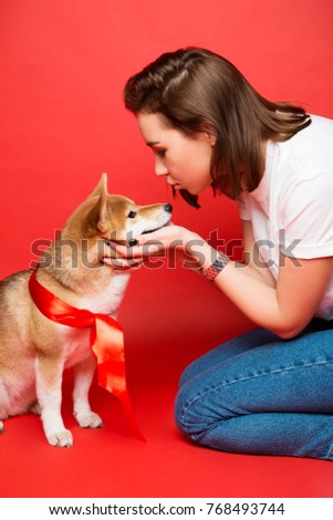 full portrait of young brunette woman in white t shirt, jeans and white sneakers sitting with a yellow white dog with red bow on  plain red background