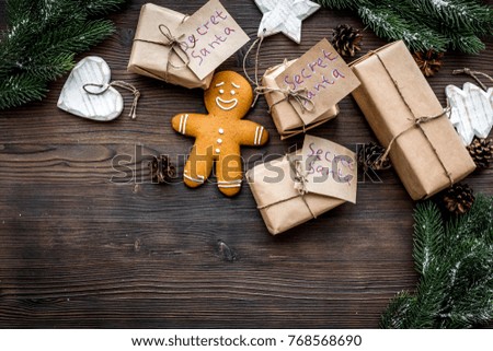 Gift exchange. Boxes with note Santa Secret near spruce branch and gingerbread cookies on dark wooden background top view copyspace