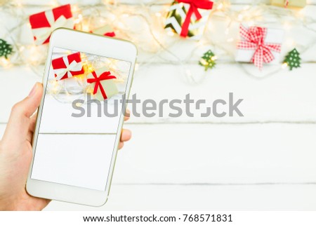 Close up hand holding mobile smartphone take Christmas New year theme red and craft gift box ribbon bow with sparkling light on white wooden table background with copy space.Displaying picture.