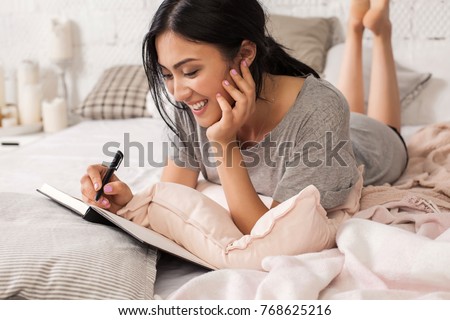 Beautiful young asian woman laying on bed and writing a diary. Smiling brunette lady with notebook and pen in her hands. Modern bright home interior on background. Royalty-Free Stock Photo #768625216