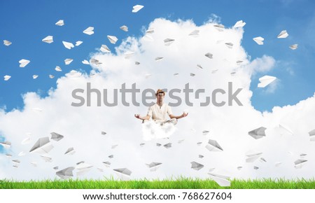 Young man keeping eyes closed and looking concentrated while meditating on cloud among flying paper planes with bright and beautiful landscape on background.