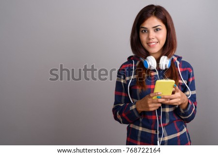 Studio shot of young beautiful Indian woman wearing headphones and using mobile phone against gray background