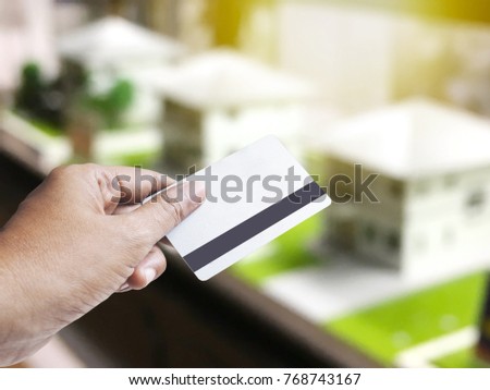 Hand holding credit card over abstract blurred background of house model samples on shelves for real estate and architecture business concept. Montage style.